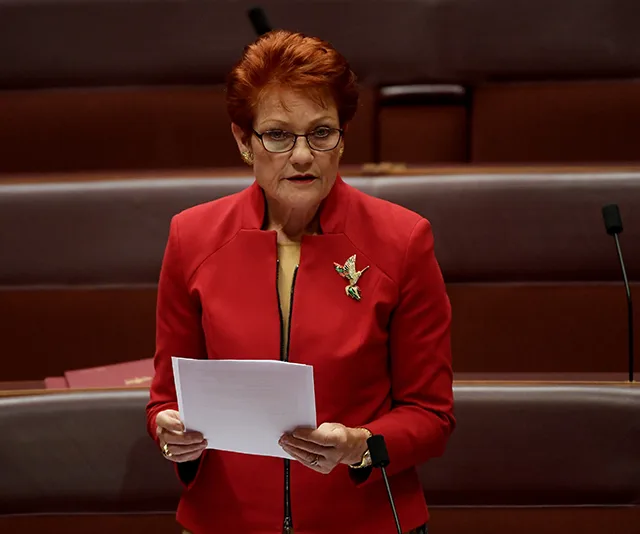 A person in a red jacket speaks in a legislative chamber, holding papers and wearing glasses.