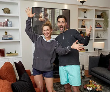 Two people joyfully pose in a well-decorated lounge with a painting, bookshelves, and comfortable seating.