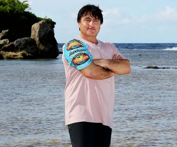 Man in a pink shirt with crossed arms, standing on a beach with ocean and rock in the background, wearing a Survivor buff.