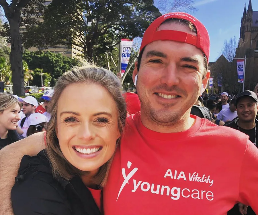 A smiling couple in matching red Youngcare shirts at an outdoor event with a crowd in the background.