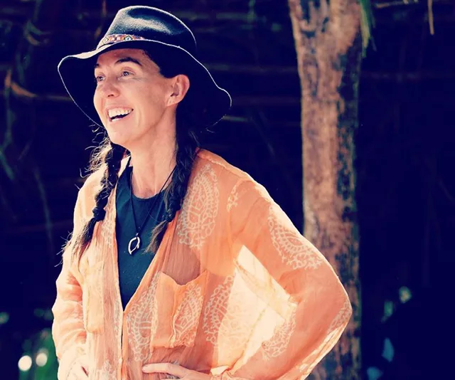 Smiling woman in a hat and flowing shirt, standing outdoors near a tree.