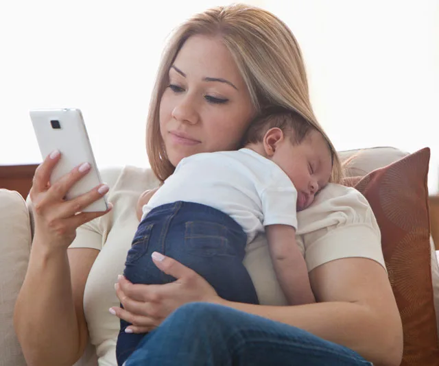 Mother multitasks by using a smartphone while holding a sleeping baby on the couch.