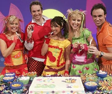 A group of five people celebrate with a large cake and colorful outfits, appearing to be from a children's TV show.