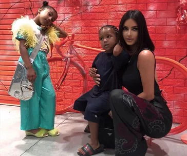 Kim Kardashian poses with two children against a vibrant red wall with a bicycle design.
