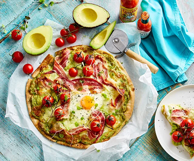 Avocado pizza topped with cherry tomatoes, bacon, and a sunny-side-up egg, served on parchment with hot sauce and fresh avocados.