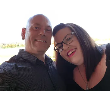 A smiling couple taking a selfie outdoors with a bright, clear sky background.