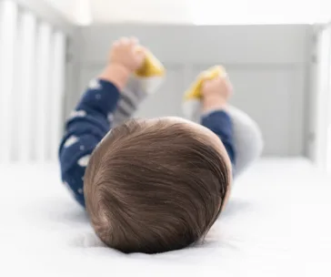 Baby lying in crib wearing navy pajamas, holding yellow toys with feet, and looking upwards.
