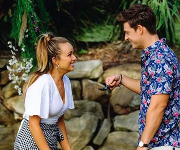 Two people smiling and talking outdoors near a stone wall and greenery.