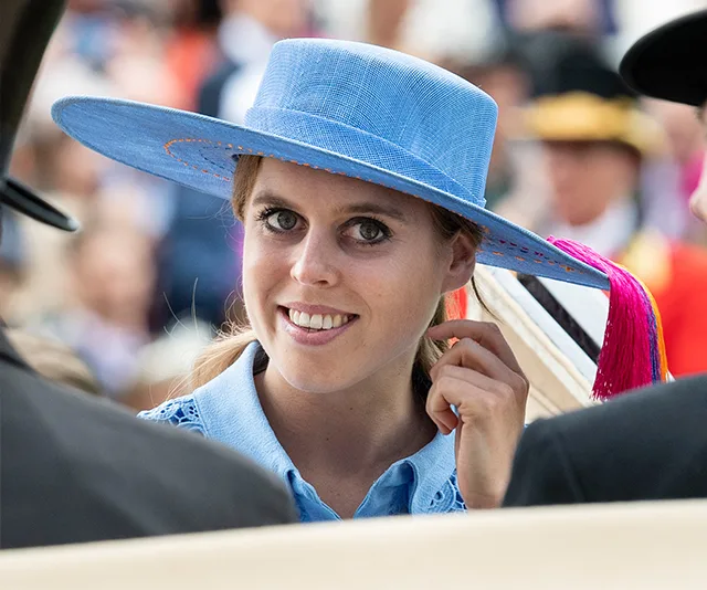 Smiling woman in a blue hat at a formal event, surrounded by people in hats.