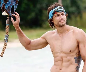 A shirtless man with a tribal tattoo and bandana stands outdoors, holding a wooden structure, set in nature.
