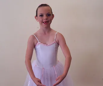 Young girl in a ballet costume poses, smiling, with hair tied back and arms gracefully positioned, in a performance setting.
