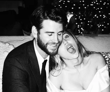 A black and white photo of a couple sitting together on a couch, the woman playfully leaning on the man's shoulder.