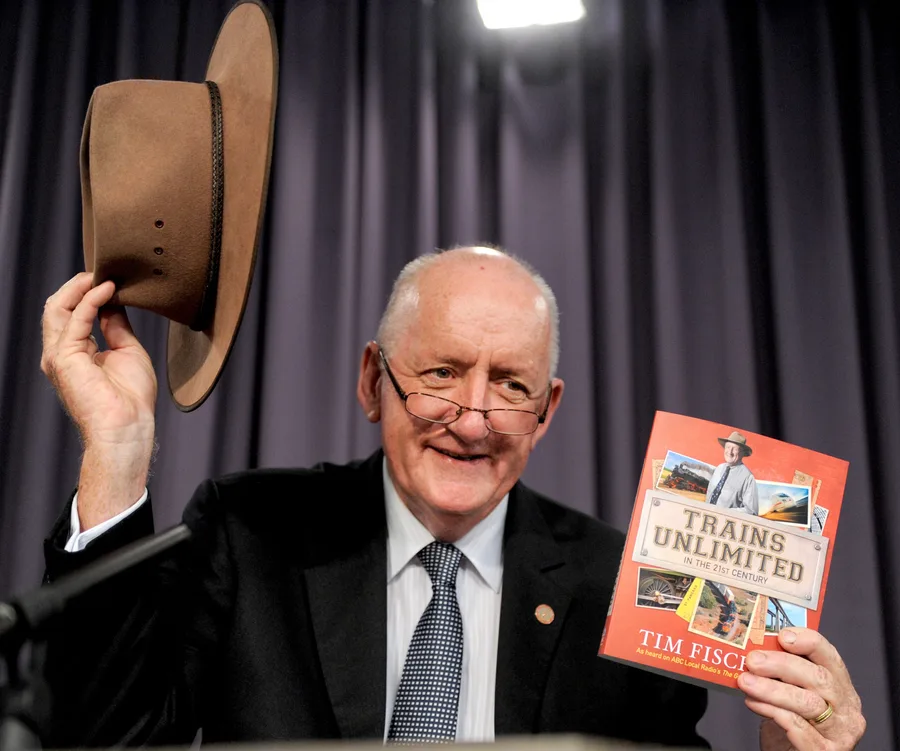 Man holding a hat and a book titled "Trains Unlimited" by Tim Fischer, standing in front of a curtain.