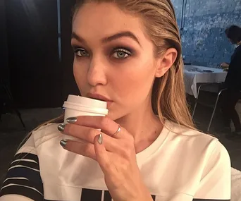 A person with long hair, wearing a white shirt, holding a small cup close to their face.