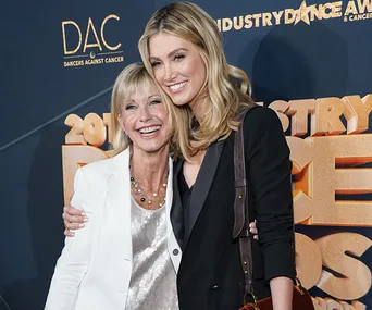 Two women smiling and embracing at an event with a blue and gold background promoting dance and cancer awareness.