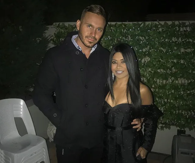 A couple in formal attire poses at night near greenery and white chairs.