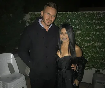 A couple in formal attire poses at night near greenery and white chairs.