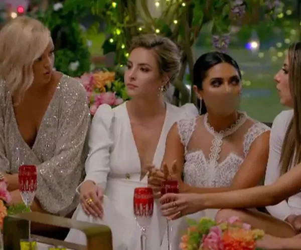 Women in evening dresses sitting together at a decorated table with drinks in red glasses, engaged in conversation.
