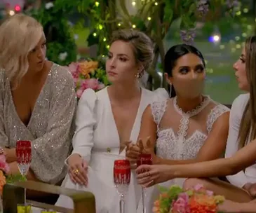 Women in evening dresses sitting together at a decorated table with drinks in red glasses, engaged in conversation.