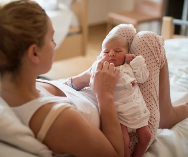 Mother in pajamas cradling a newborn baby on her legs, in a cozy room setting.