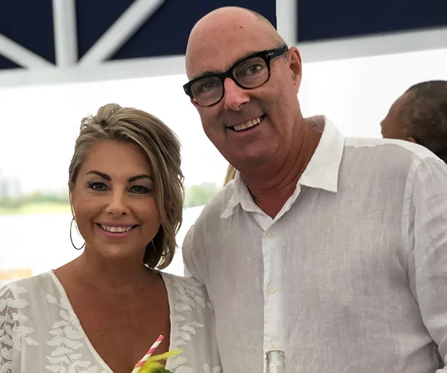 Man and woman smiling together at an outdoor event, both wearing white clothing.