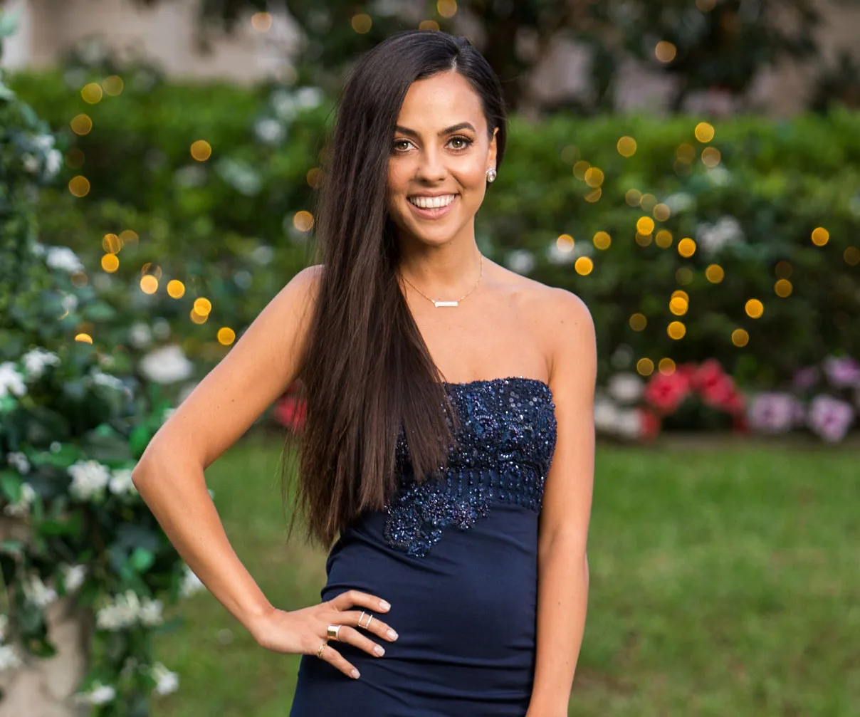 Woman in a beaded navy dress smiles outdoors with blurred lights and greenery in background.
