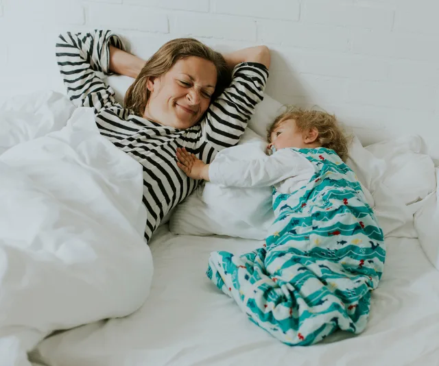Woman in striped shirt smiling at baby in a colorful sleeping bag on bed.