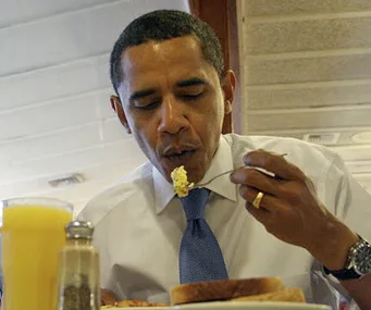A man in a white shirt and blue tie eating scrambled eggs, next to orange juice and a salt shaker.