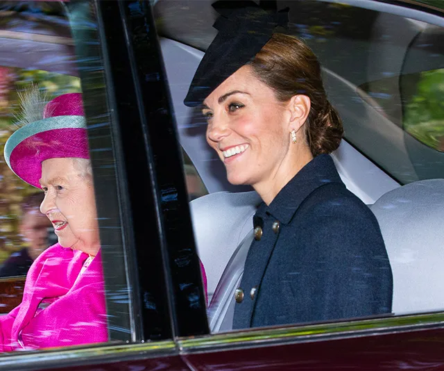 Two women in a car, one in a pink outfit with a hat, and one in a dark coat with a black hat, smiling.