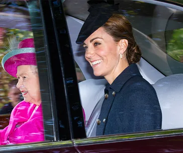 Two women in a car, one in a pink outfit with a hat, and one in a dark coat with a black hat, smiling.