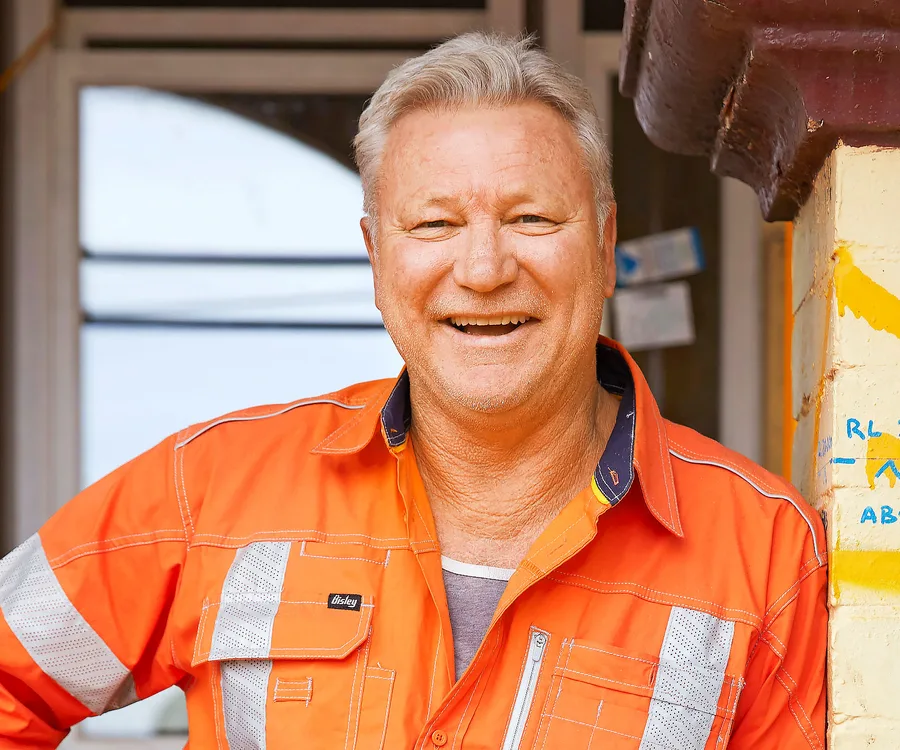 An individual in an orange work shirt smiles, standing beside a painted wall.