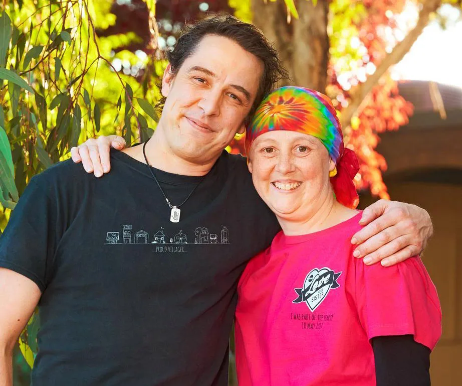 A person in a black shirt hugs another in a tie-dye bandana and pink "Love Your Sister" shirt, smiling outdoors.