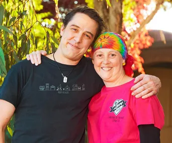 A person in a black shirt hugs another in a tie-dye bandana and pink "Love Your Sister" shirt, smiling outdoors.