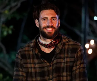 A man with a beard in a plaid shirt smiles at night, wearing a Survivor-themed buff around his neck.