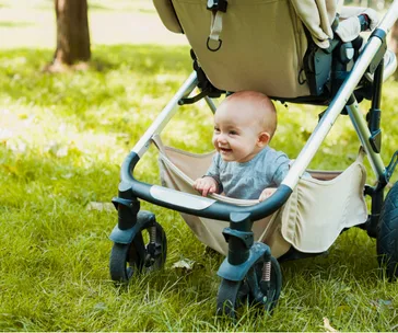 A baby smiling while sitting in a beige pram on a grassy lawn in the park.