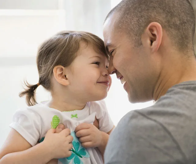 Father and daughter smiling, touching noses, embracing warmly indoors.