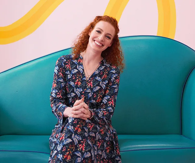 A person with curly red hair in a floral dress sits smiling on a turquoise couch against a pastel-patterned wall.