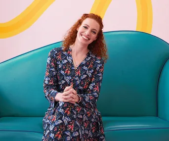 A person with curly red hair in a floral dress sits smiling on a turquoise couch against a pastel-patterned wall.