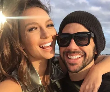 A smiling woman and man wearing sunglasses and a beanie, embracing outdoors with a cloudy sky background.