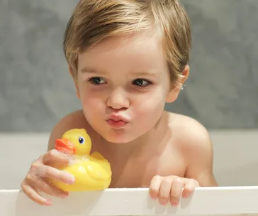 Young child in a bathtub holding a yellow rubber duck, making a playful face with closed lips.