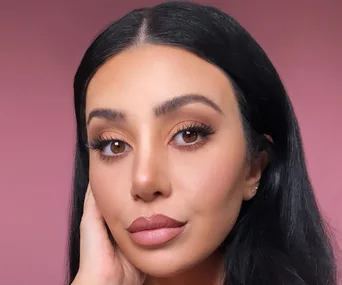 A woman with long black hair and makeup poses against a pink background.