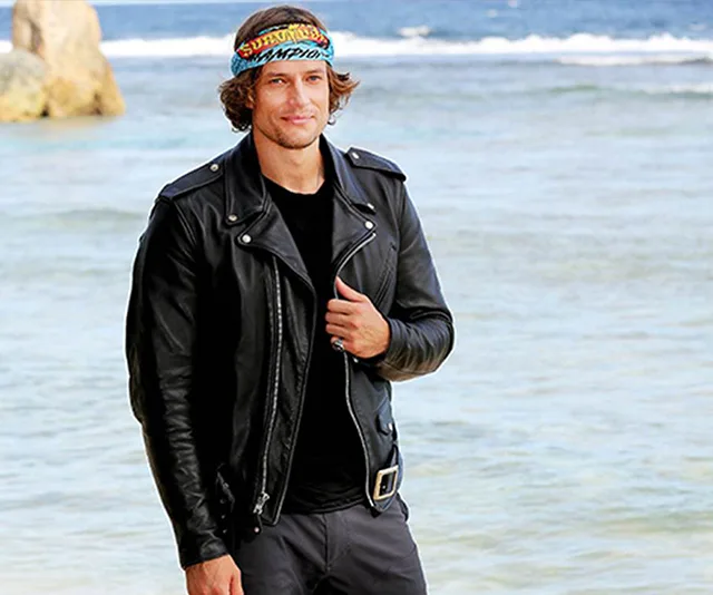Man in leather jacket and bandana stands on a beach, smiling with ocean in the background.