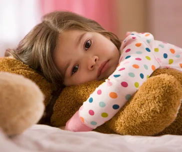A young child in polka dot pajamas hugs a teddy bear, looking sad while lying on a bed.