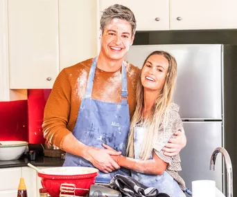 A smiling couple in a kitchen wearing aprons, covered in flour from baking, standing in front of a fridge.