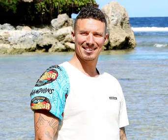 A man in a white shirt stands on a beach with clear blue water and rocky background, wearing a colorful arm sleeve.