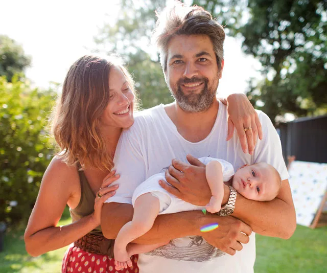 A happy couple with a newborn baby in the father's arms, smiling outdoors in a garden setting.