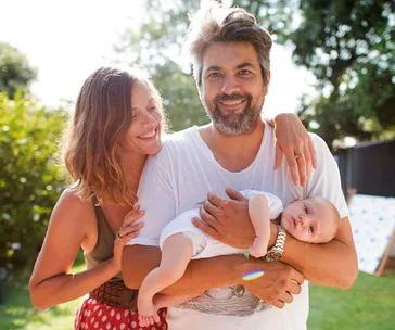 A happy couple with a newborn baby in the father's arms, smiling outdoors in a garden setting.