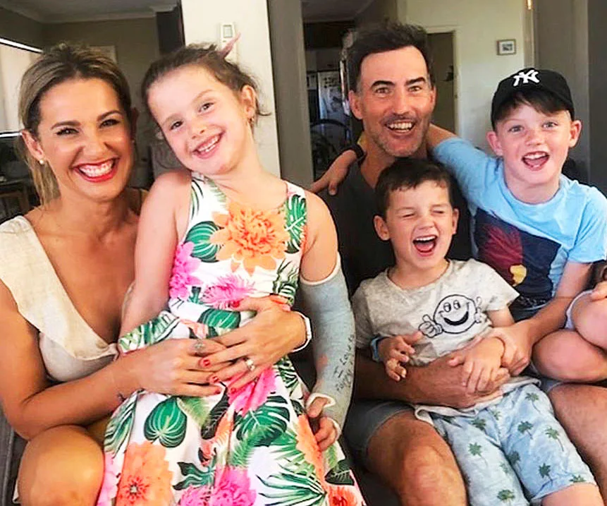 Family smiling and sitting on a couch; two adults and three children. One child wears a floral dress with a cast.