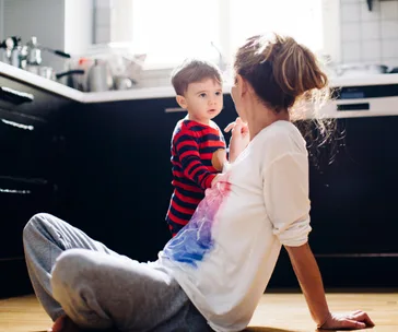 A mother sits on the kitchen floor, engaging with her toddler who is standing and wearing a red striped shirt.