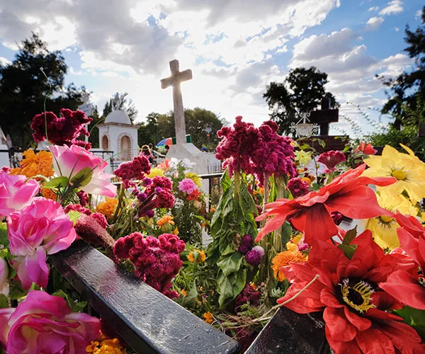 A cemetery adorned with colorful flowers and a prominent cross against a partly cloudy sky.